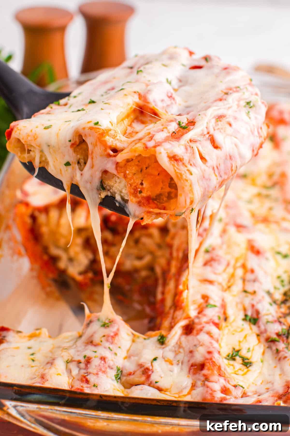 Classic Baked Manicotti with Ricotta and Marinara 2 A close-up of a spatula lifting two stuffed manicotti from a baking dish, with stretchy melted cheese and a visible creamy filling inside the pasta. The dish is surrounded by more baked pasta and cheese.