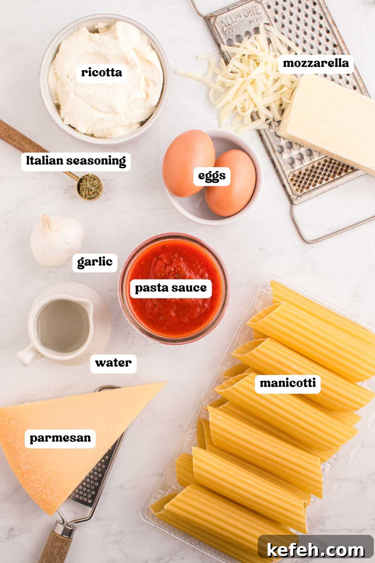 Classic Baked Manicotti with Ricotta and Marinara 4 Overhead image showing ingredients for baked manicotti, each labeled: ricotta in a bowl, shredded mozzarella on a grater, eggs in a small bowl, Italian seasoning in a spoon, garlic, pasta sauce in a glass bowl, a small pitcher of water, a wedge of parmesan with a grater, and uncooked manicotti in a plastic tray.