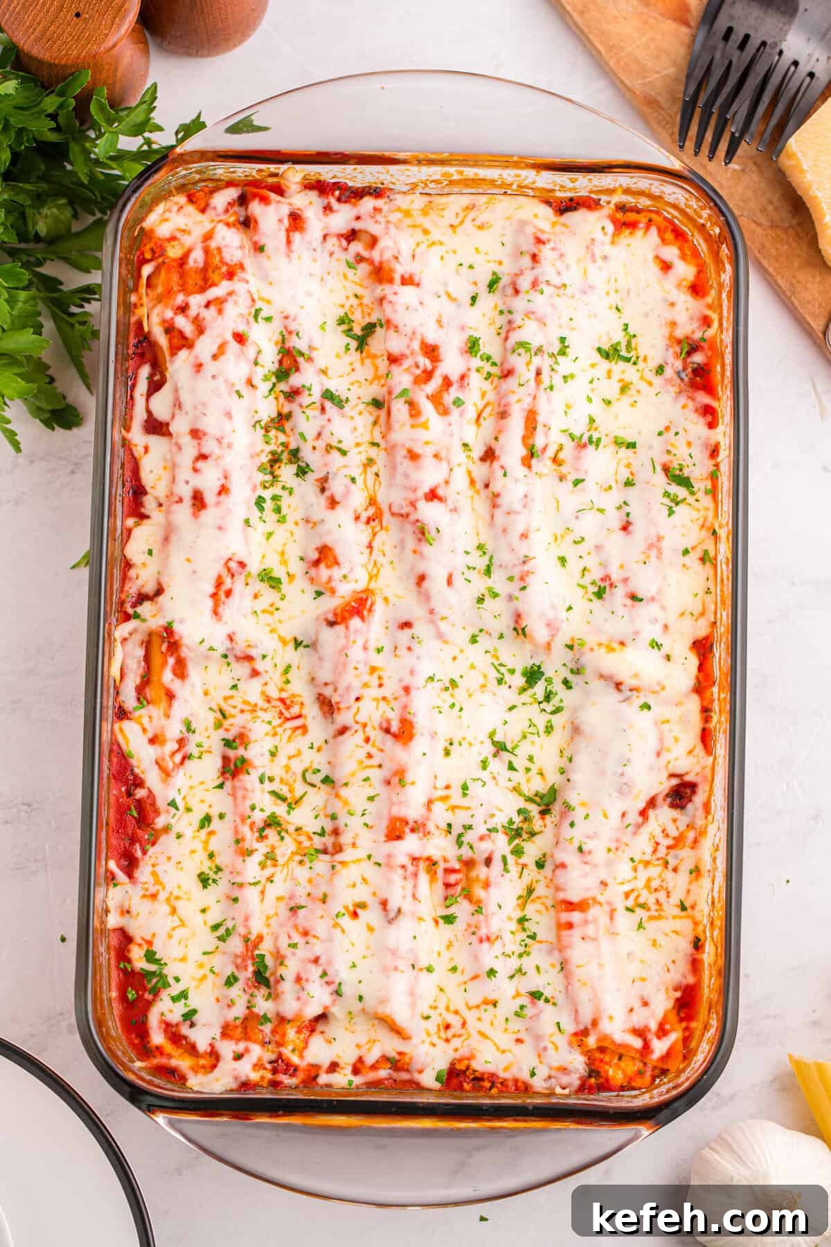 Classic Baked Manicotti with Ricotta and Marinara 10 Overhead shot of a glass baking dish filled with neatly arranged manicotti covered in melted mozzarella and tomato sauce, garnished with chopped parsley.