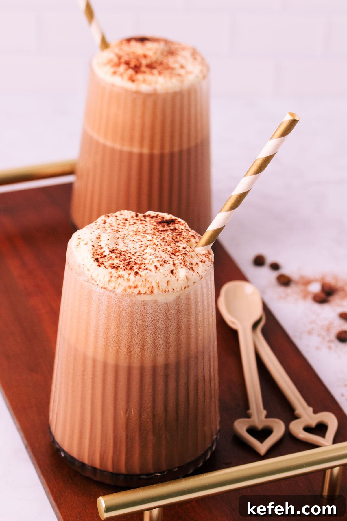 Creamy Mocha Milkshake Recipe for Coffee Lovers 2 Two mocha milkshakes in glasses with a ribbed texture, topped with whipped cream and a sprinkle of cocoa, with gold and white striped paper straws on a dark wooden board with gold handles.