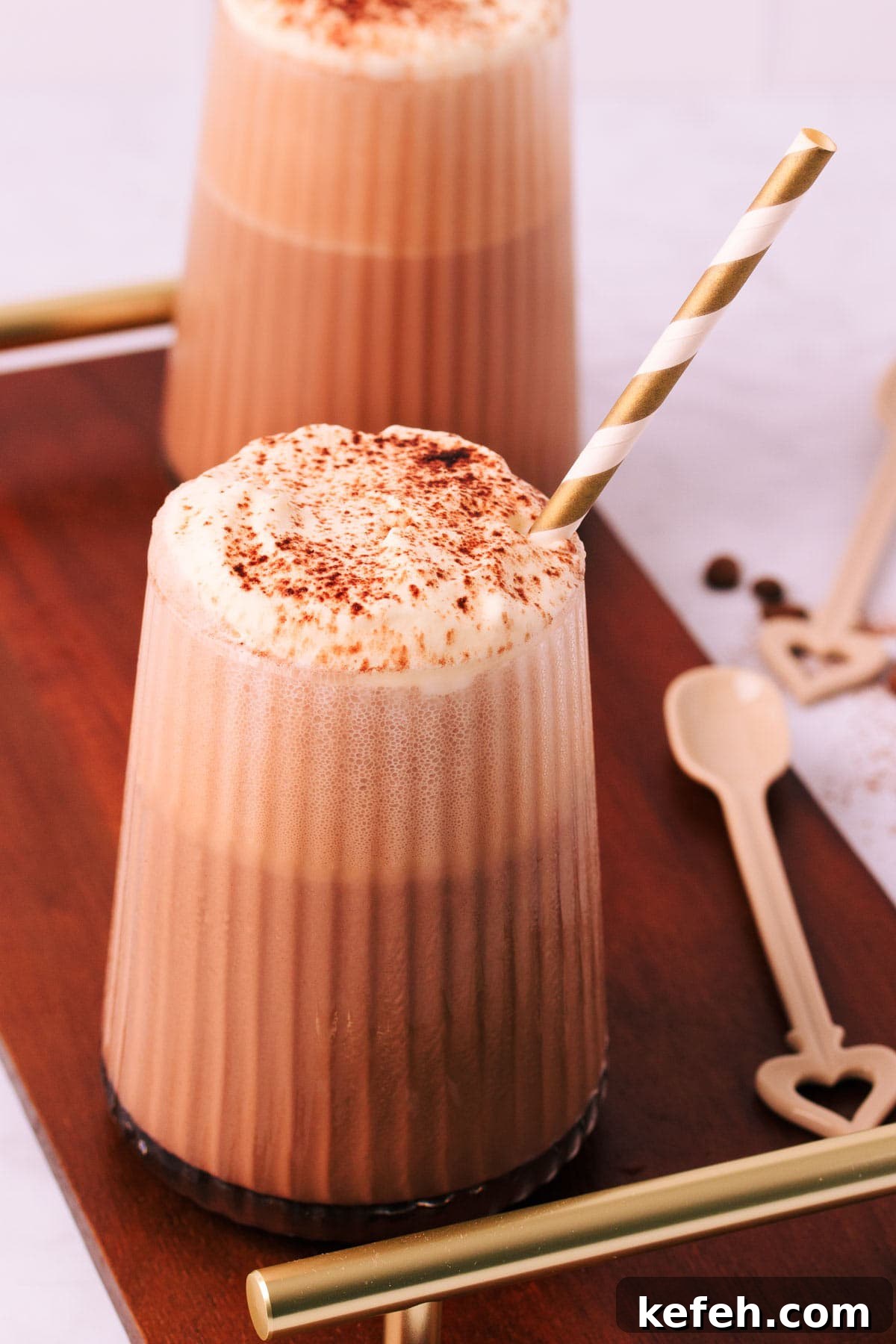 Creamy Mocha Milkshake Recipe for Coffee Lovers 3 A mocha milkshake in a tapered glass with a ribbed texture, topped with whipped cream and a sprinkle of cocoa, with a gold and white striped paper straw on a dark wooden board with gold handles and a mocha-coloured ceramic spoon.