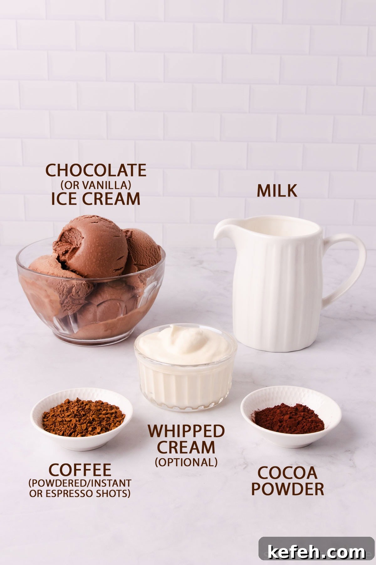 Creamy Mocha Milkshake Recipe for Coffee Lovers 4 The mocha milkshake ingredients - chocolate ice cream in a glass bowl, milk in a white jug, coffee, cocoa powder and whipped cream in small bowls, on a pale grey marble background.