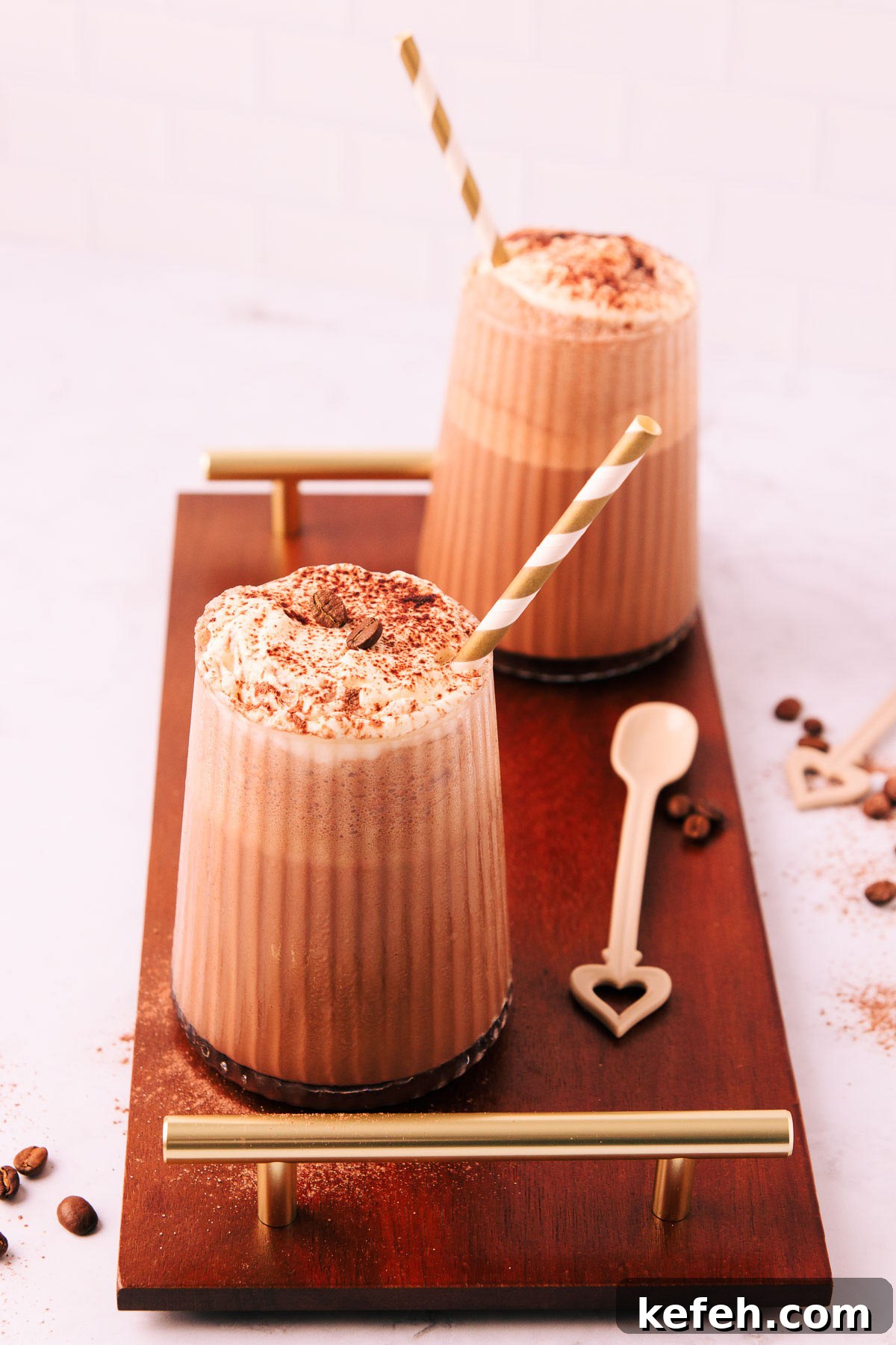 Creamy Mocha Milkshake Recipe for Coffee Lovers 7 Two mocha milkshakes in glasses with a ribbed texture, topped with whipped cream and a sprinkle of cocoa, with gold and white striped paper straws on a dark wooden board with gold handles.