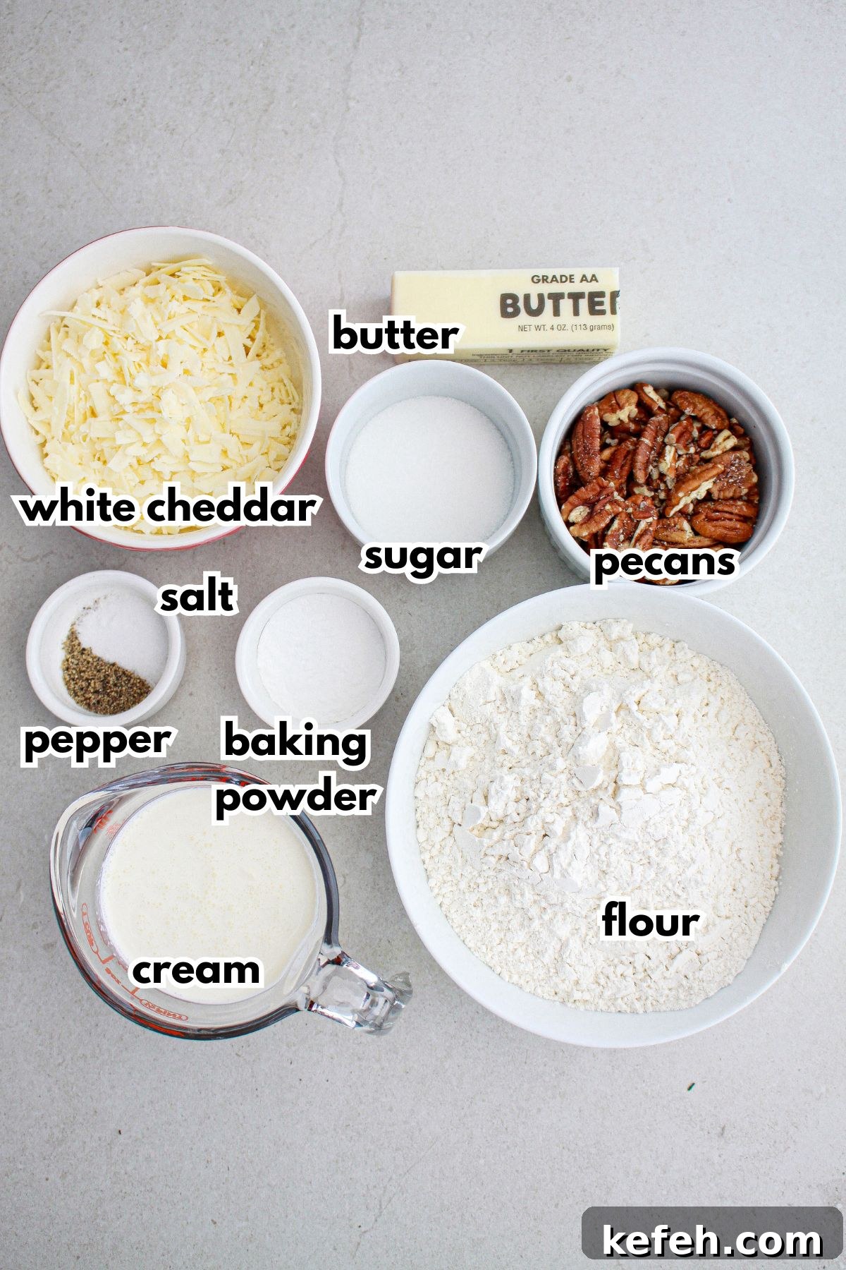bowls of white cheddar cheese, butter, sugar, pecans, salt, pepper, baking powder, flour, and cream on a counter.