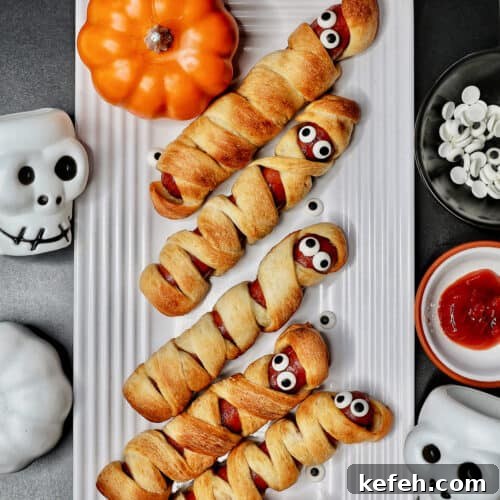 mummy hot dogs on a white plate surrounded by Halloween decorations like mini pumpkins and skull bowls.