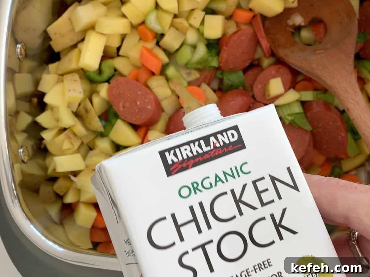 Pouring chicken stock into a slow cooker with chopped vegetables and sausage.