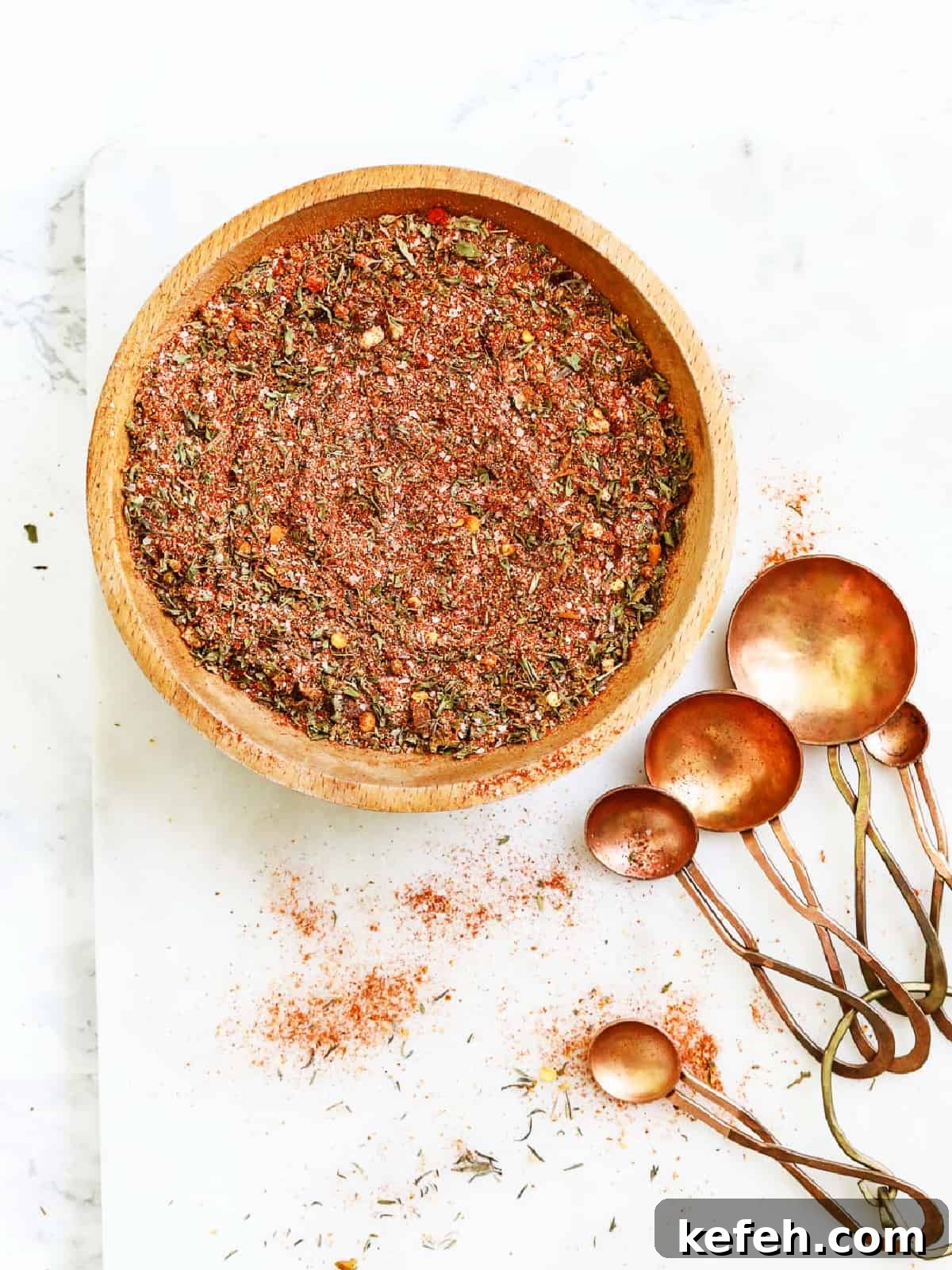 A wooden bowl with mixed Cajun seasoning and copper measuring spoons on a marble countertop.