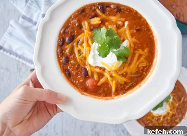 Turkey Taco Chili Soup Recipe — Hearty Weeknight Meal 6 hand picking up bowl of turkey chili with yogurt, cheese, cilantro on top.