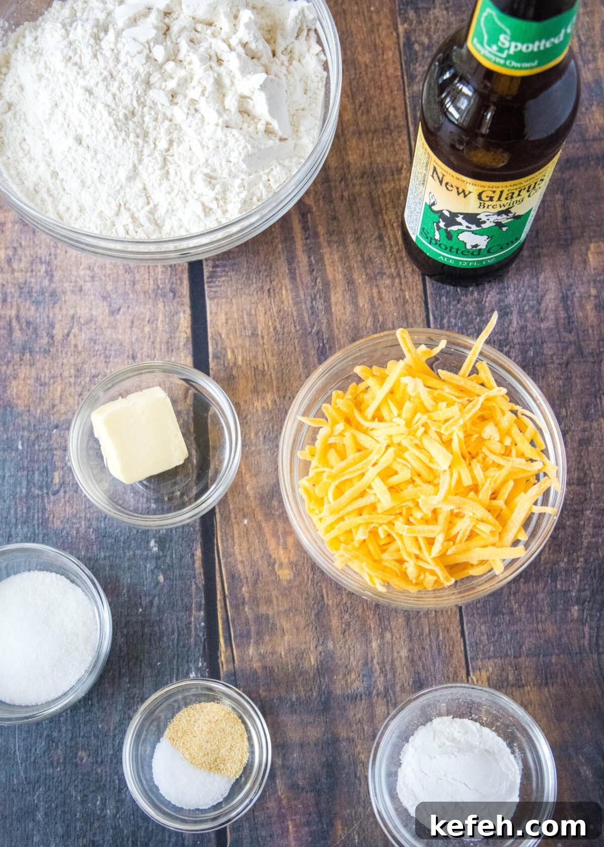 This easy cheddar beer bread recipe is moist, soft, full of beer and cheese flavors, and so quick and easy to make at home. Overhead view of the ingredients needed for beer bread: a bowl of flour, a bowl of cheese, a bowl of baking powder, a bowl of sugar, a bowl of salt and garlic powder, a bowl of butter, and a bottle of beer