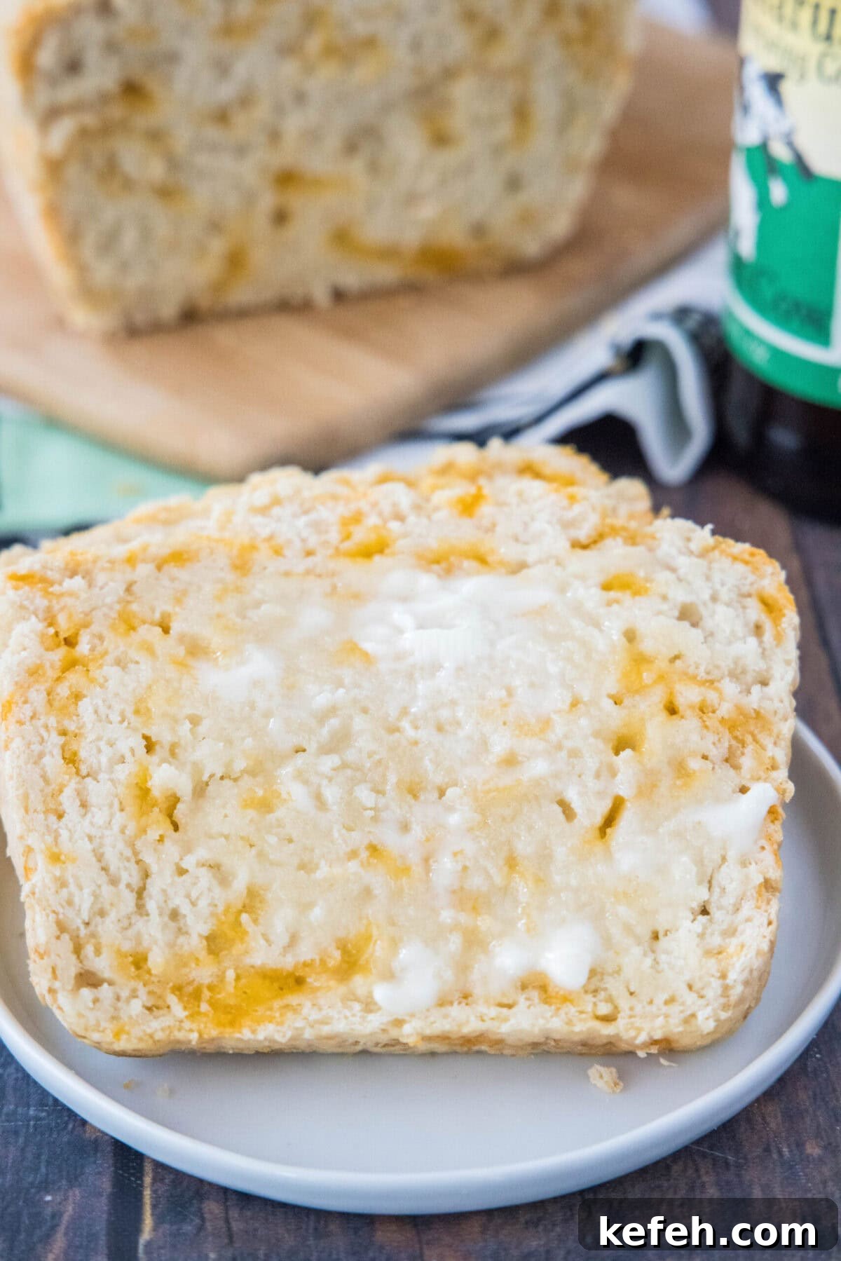 This easy cheddar beer bread recipe is moist, soft, full of beer and cheese flavors, and so quick and easy to make at home. A piece of beer bread on a plate, covered in butter, with a loaf behind it