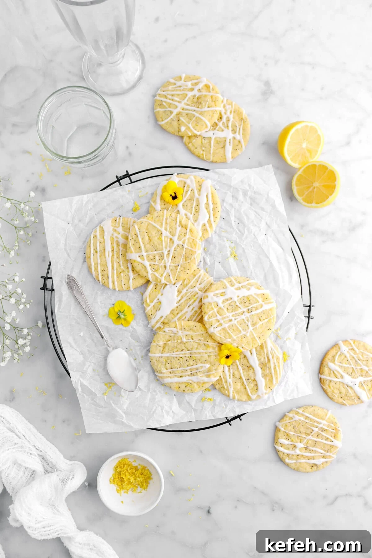 Lemon Poppy Seed Cookies with Lemon Drizzle Recipe 2 pulled back overhead shot of lemon poppy seed cookies on parchment lined cooling rack with yellow flowers on top and spoon of icing beside, with four more cookies around on marble surface, with two lemon halves and empty glasses above.